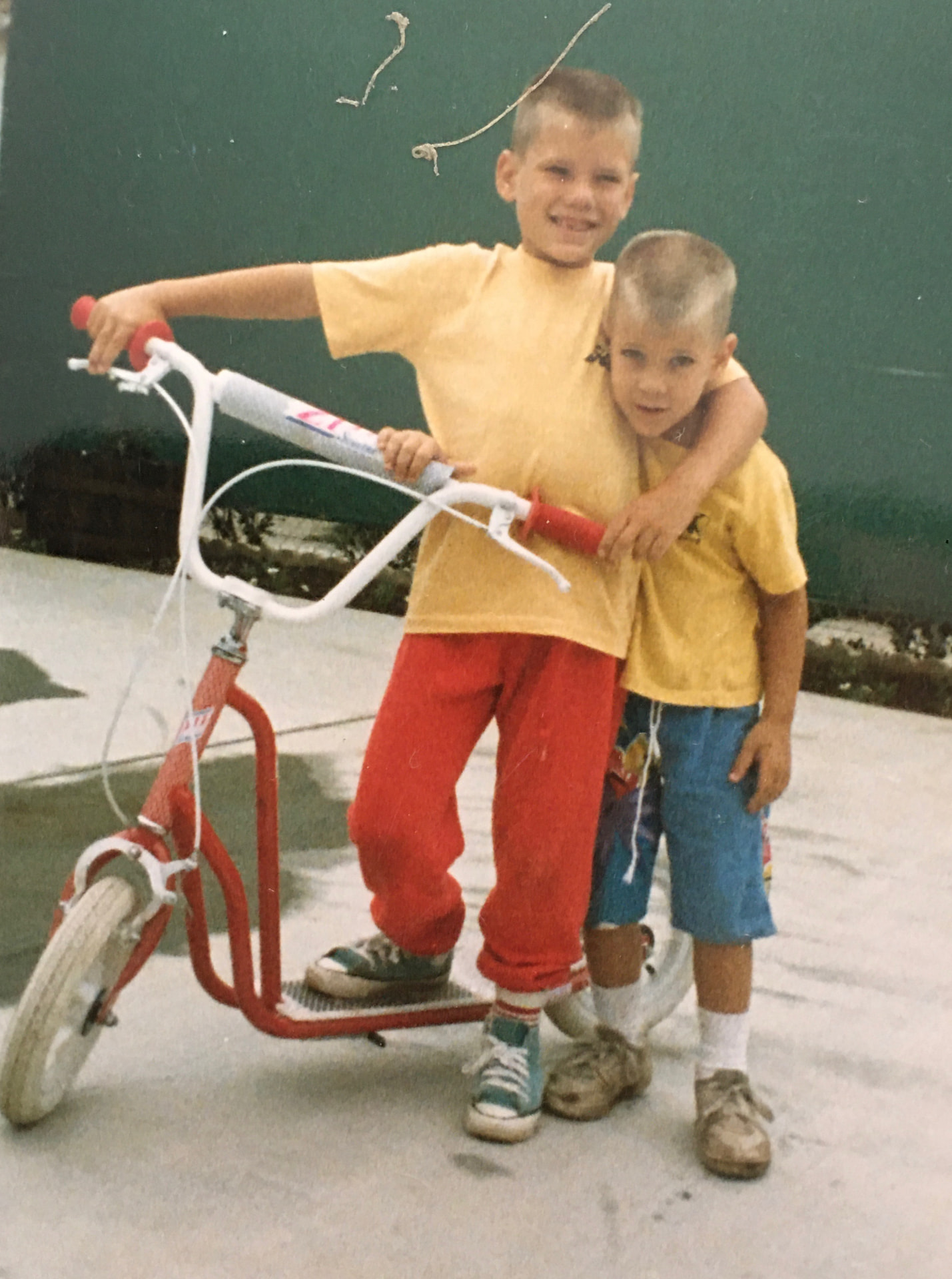 Scott and Steven as young boys, the older brother on a red scooter with his arm around his younger brother